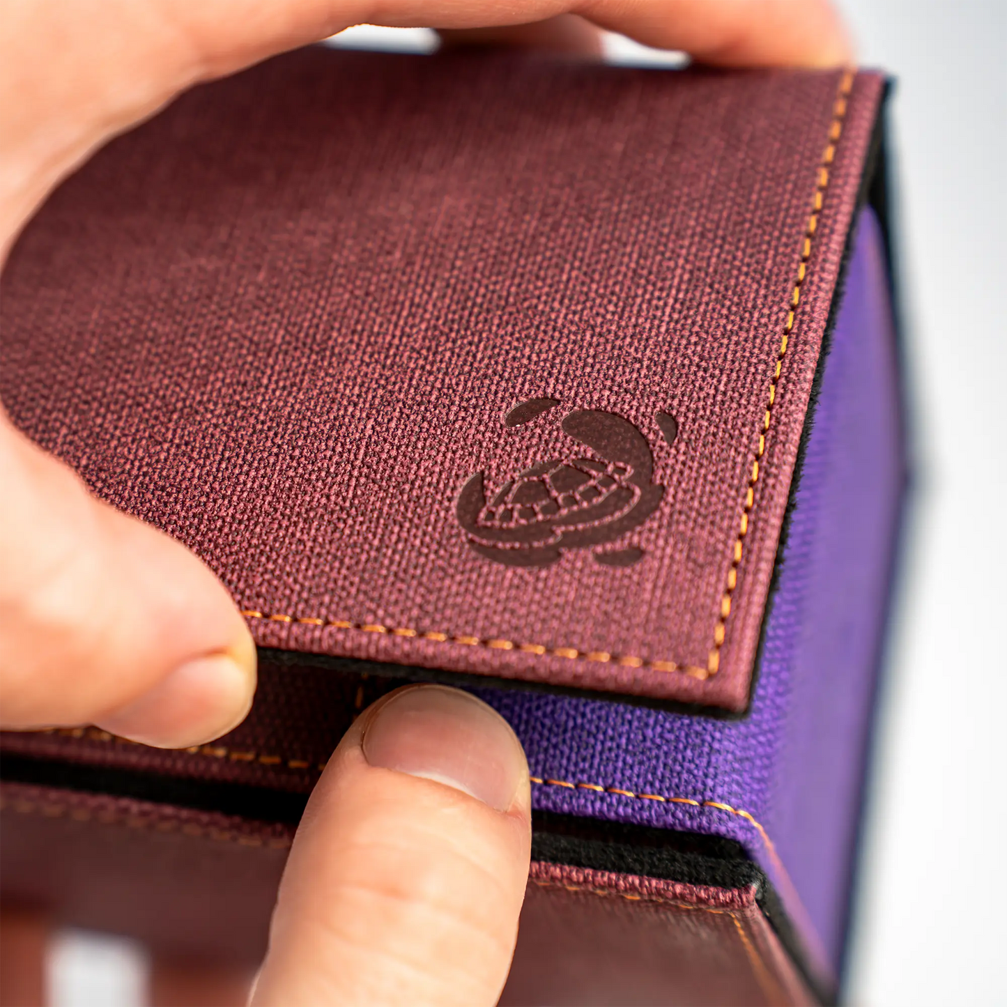 hands opening multi-color wine red, purple and black dual material resistant denim-like caratex and soft resiplush tourneycove 120+ tournament deck box for tcg trading cards, with removable, magnetic compartment for dice, tokens and counters and extra card divider